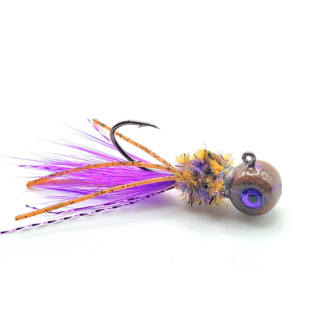 "Hand tied crappie jig with a purple feather tail, a yellow and brown sparkly body made of synthetic material, and a round brown jig head with a blue dot. This crappie lure features a sickle hook, designed to attract fish by mimicking the appearance and movement of prey in the water, making it an effective and visually appealing bait for crappie fishing. This fishing lure is hand tied by Ramble Tamble Tackle."