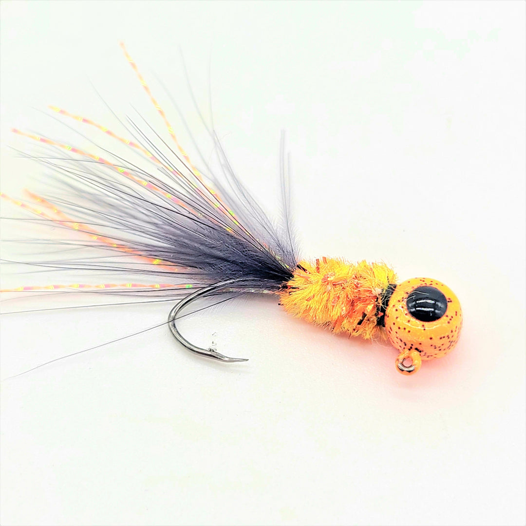"Hand tied crappie jig with a black feather tail, a bright yellow sparkly body made of synthetic material, and a round orange jig head with black speckles. This crappie lure features a sickle hook, designed to attract fish by mimicking the appearance and movement of prey in the water, making it an effective and visually appealing bait for crappie fishing. This fishing lure is hand tied by Ramble Tamble Tackle."